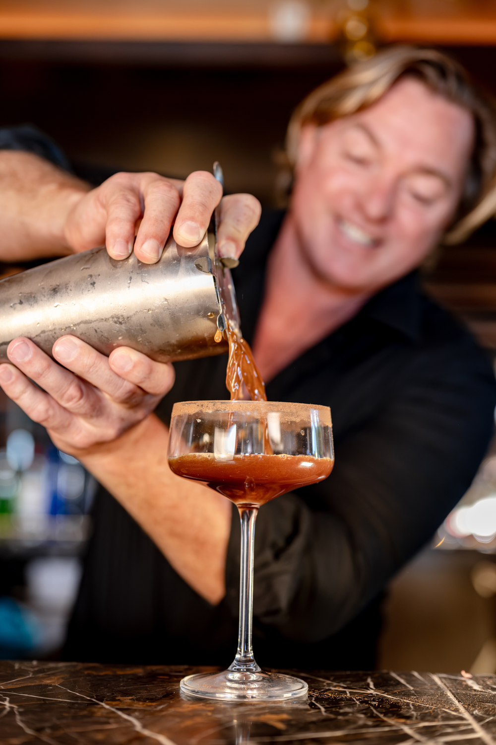 Percy pouring a cocktail from a shaker into a glass with a blurred background